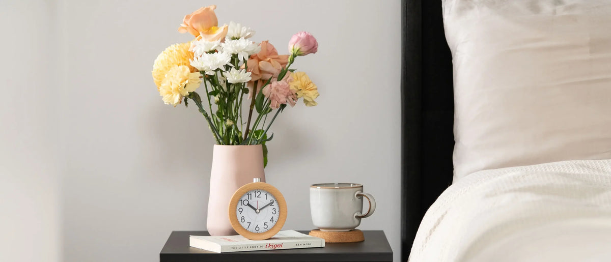 Wooden alarm clock on a bedside table with flowers and a cup of tea