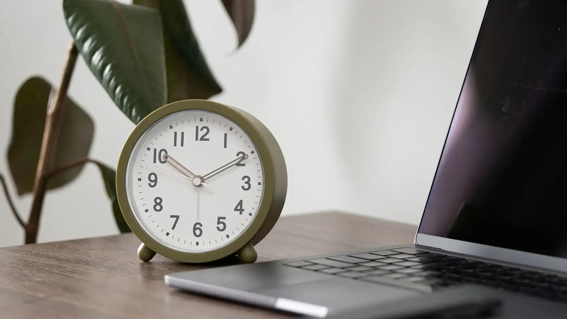 Olive green round desk alarm clock on wooden desk beside a laptop